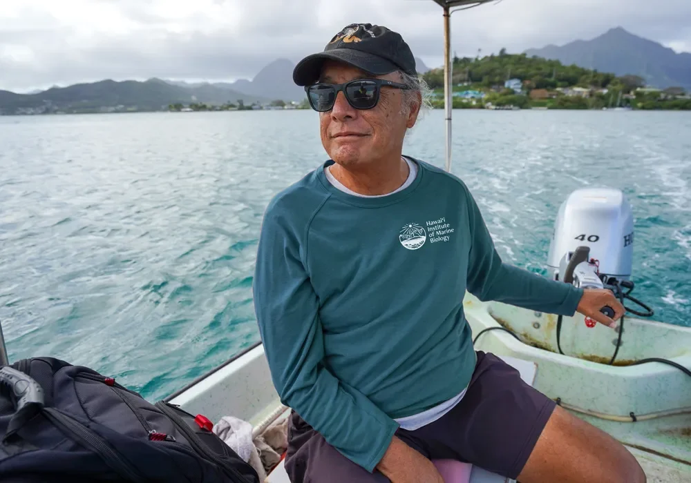 A man wearing a teal top, sunglasses and a hat steering a small boat towards an island in Hawai'i.