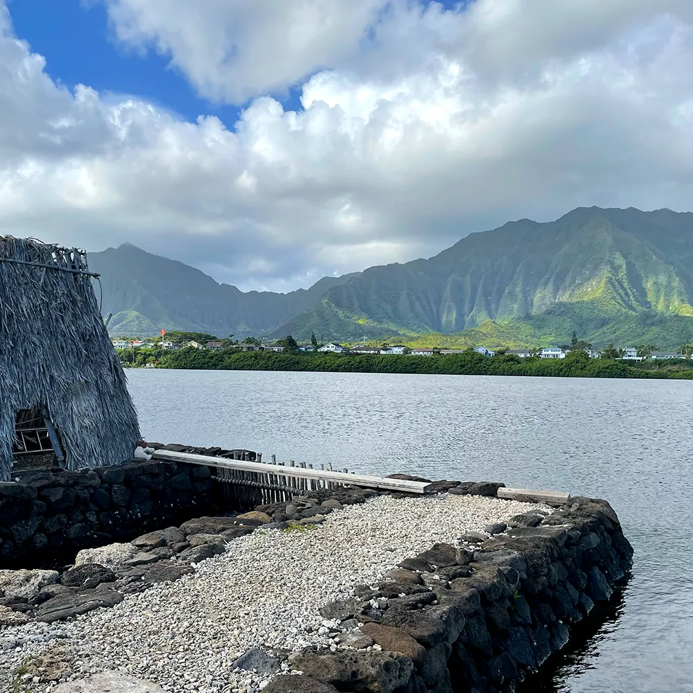 himb-splashweek-2024-fishpond A hatched structure surrounded by water and green Hawaiian mountains.