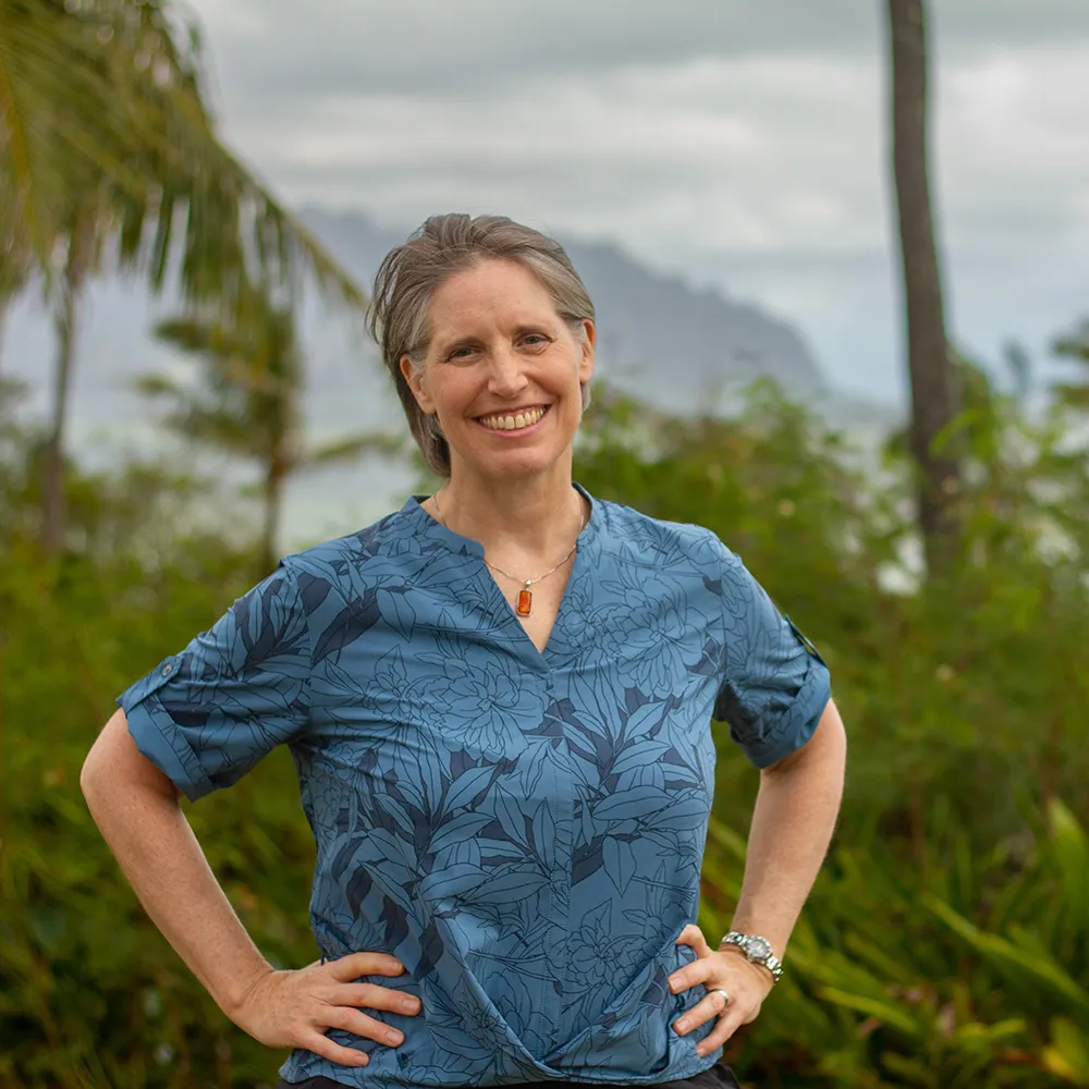 himb-splashweek-2025-megan-headshot A woman with a blue floral shirt, standing and smiling with her arms on her hips, with a backdrop of palm trees and tropical foliage.